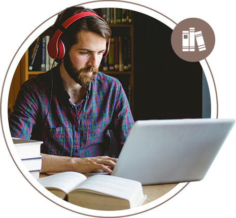 A man with brown hair and a beard wearing headphones while working on a laptop computer in a library.
