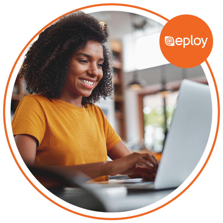 A smiling woman wearing an orange t-shirt types on a laptop in a bright, modern workspace